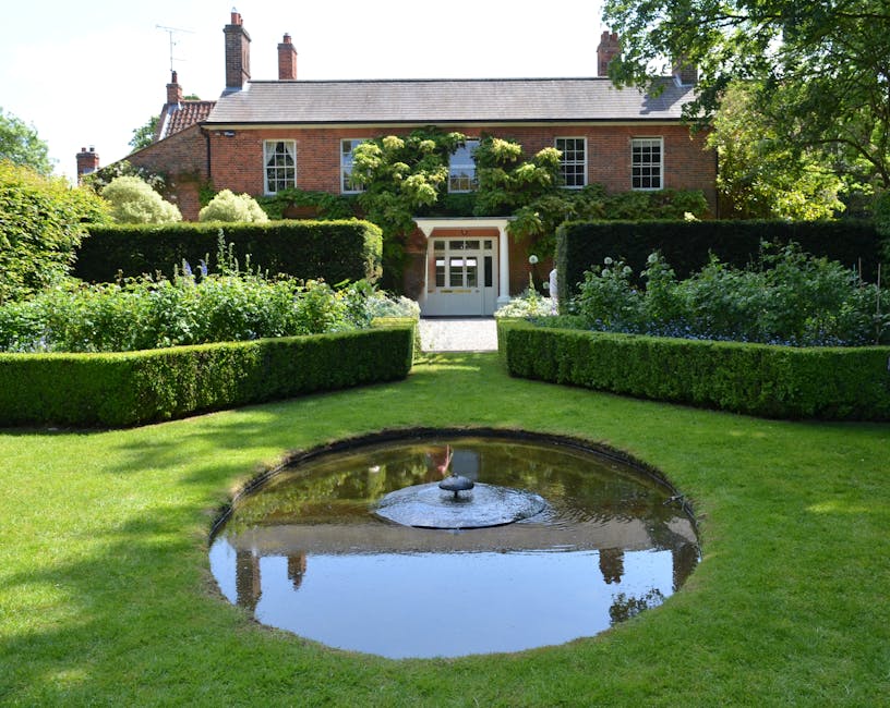 A neatly maintained garden in front of a brick house features a lush, green lawn with a small, circular pond that has a central fountain, reflecting the surrounding foliage and blue sky. Symmetrically