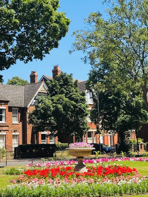 A neatly maintained garden in front of a brick house features a lush, green lawn with a small, circular pond that has a central fountain, reflecting the surrounding foliage and blue sky. Symmetrically