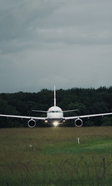 A small airplane is positioned on a grassy airfield, facing directly toward the camera with its nose slightly tilted downward. The aircraft features a white fuselage with dark windows, twin engines mo
